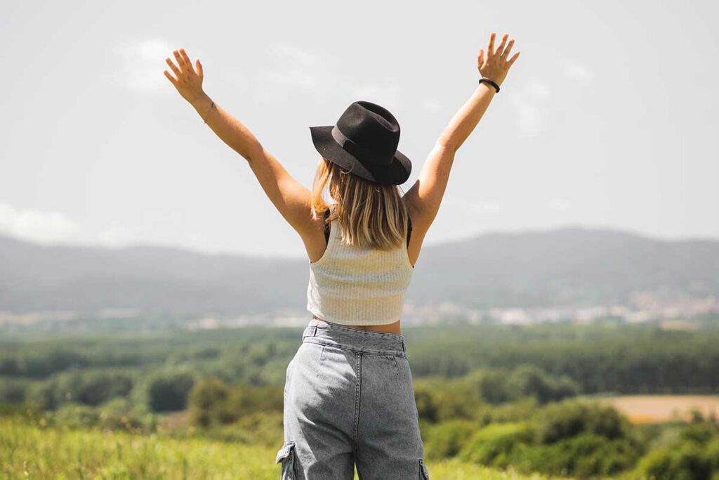 Blonde woman wearing a hat with her hands up looking towards mountains.