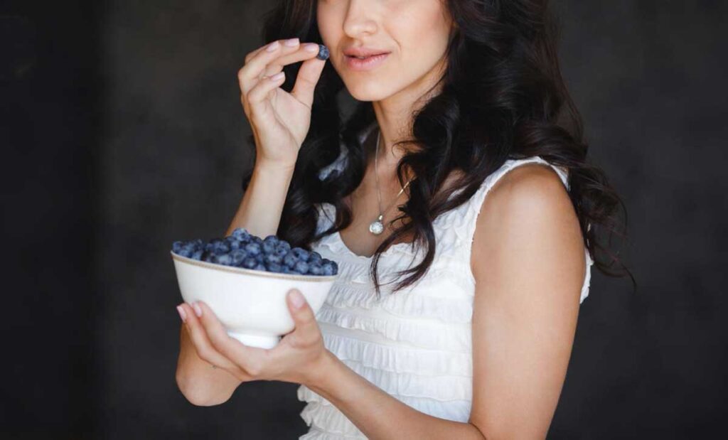 Woman in white shirt eating blueberries from a bowl.