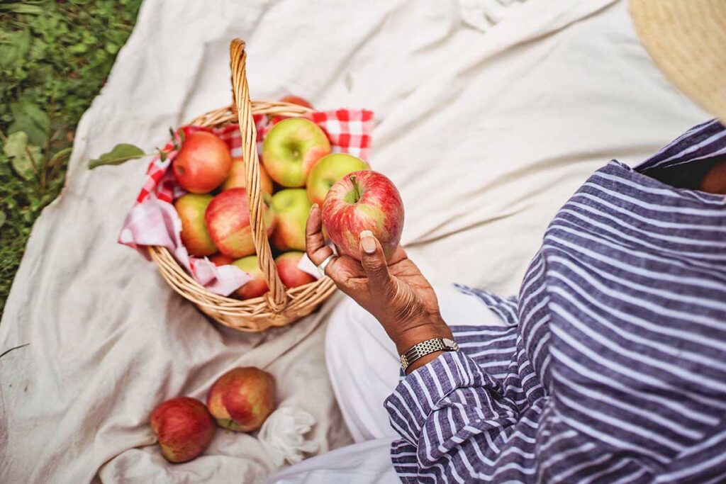 Woman sitting on a blanket with a basket of apples holding an apple.