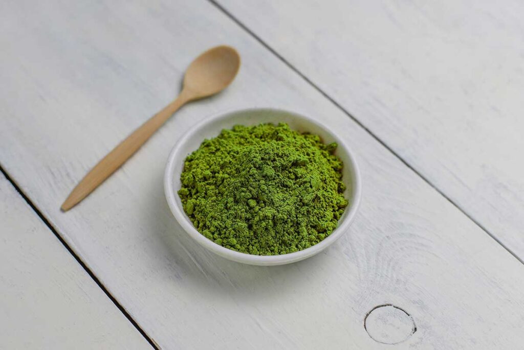 Barley grass juice powder in white bowl with wooden spoon.
