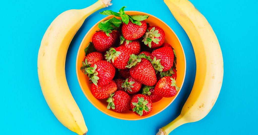 Bowl of strawberries between two bananas against blue background.