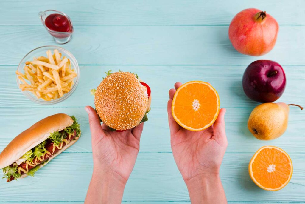 Two hands on blue background holding food along with food on the table. Junk food on the left side and fruit on the right.