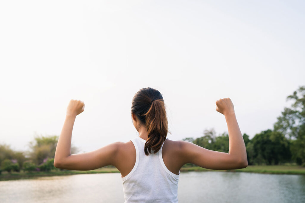 Healthy young woman holding arms up facing away toward pond.