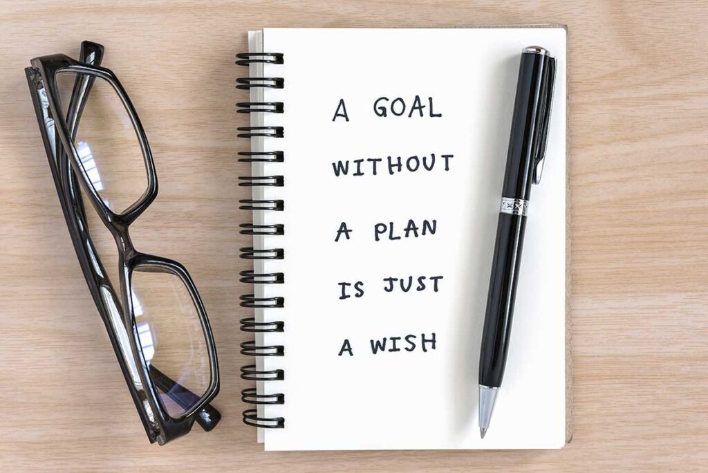 Glasses, pen, and notebook that says "A Goal Without A Plan Is Just A Wish"