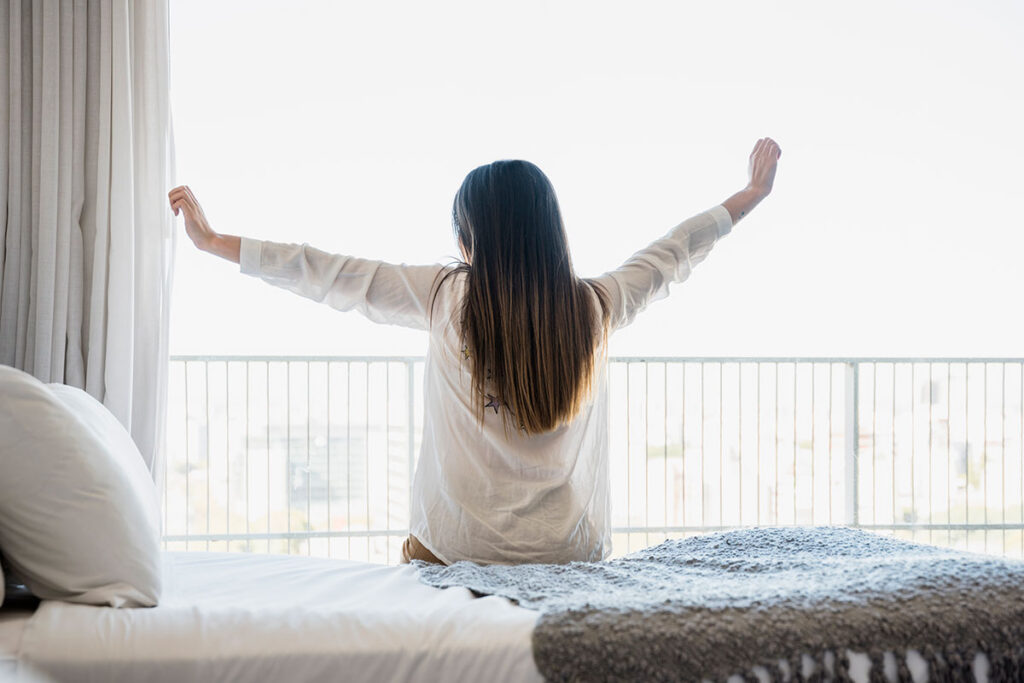 Rear view of woman sitting on bed stretching her arms.