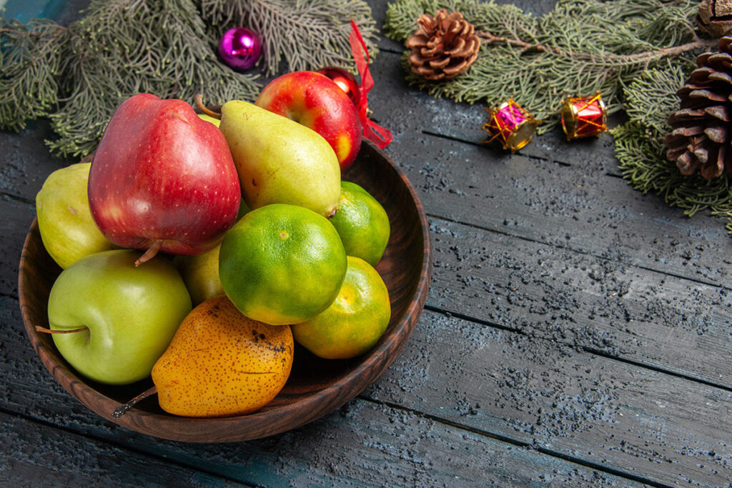 Pears, apples, and limes in a wooden bowl on wooden table with pine and Christmas ornaments. 