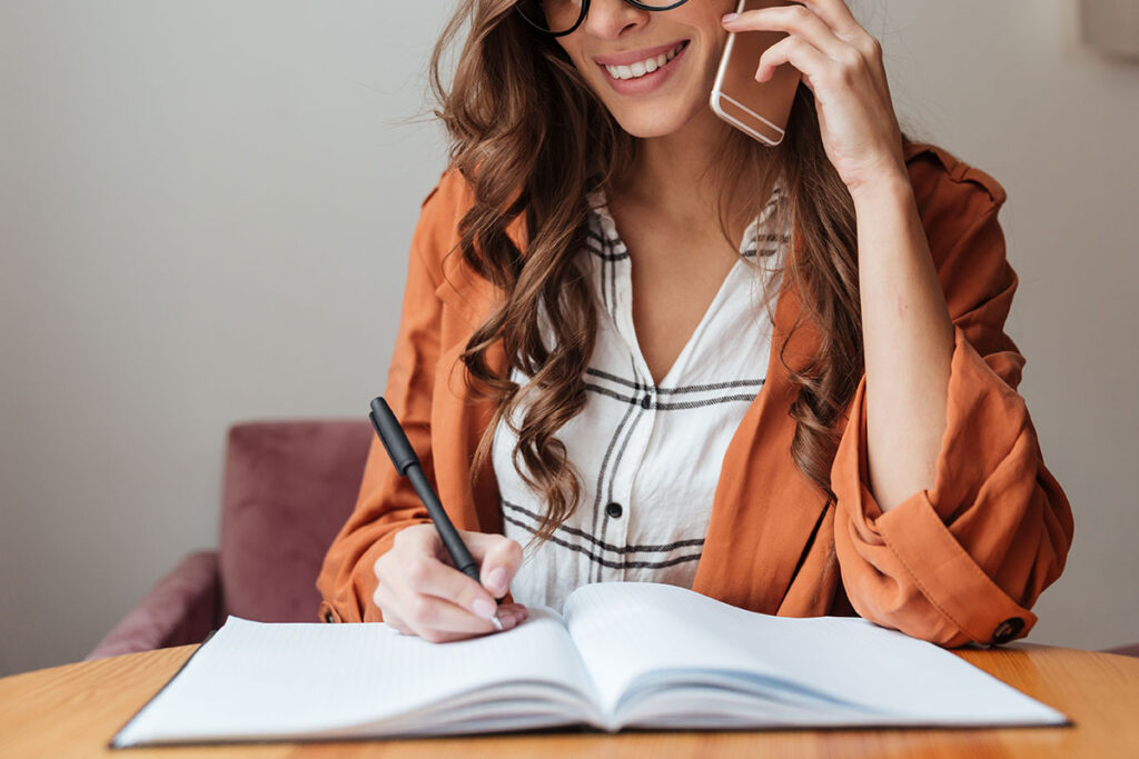 Woman with long hair and orange shirt on phone taking notes.