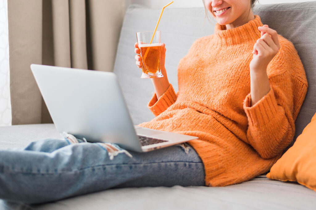 Smiling young woman in orange sweater holding drink with laptop on her lap.