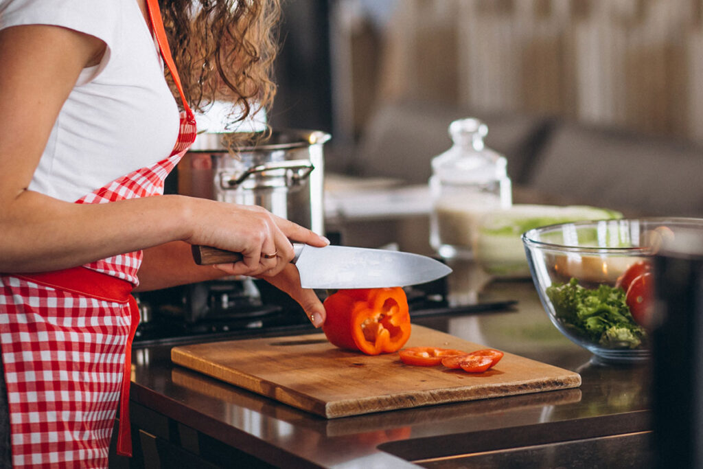 Woman in red and white checker apron chopping a red bell pepper on a cutting board.
